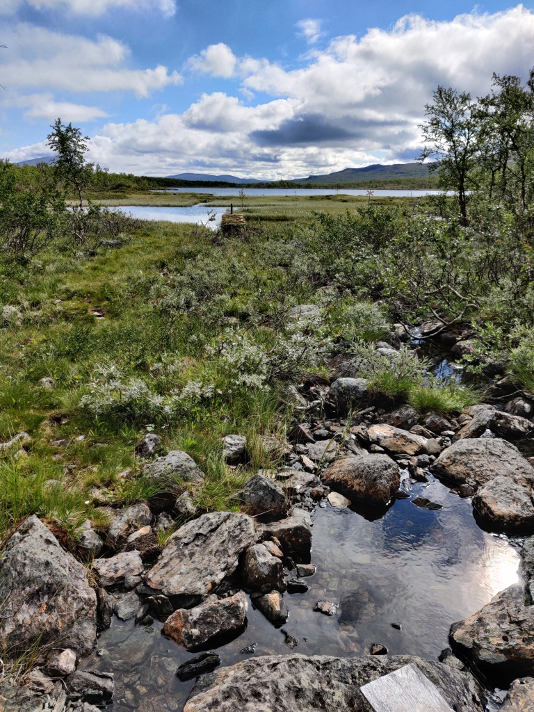 Kuva karusta maisemasta, jossa kiviä, vettä ja varpuja. Keskellä kuvaa näkyy keltaiseksi värjätty merkki, joka erottaa Suomen ja Ruotsin valtioiden rajat toisistaan.