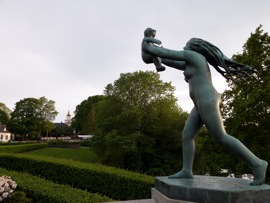 Kuva puistosta, jonka pensasaita on leikattu tarkan kulmikkaaksi. Etualalla patsas alastomasta naisesta, joka pitelee pientä lasta korkealla ilmassa. Nainen näyttää olevan liikkeessä ja hänen pitkä tukkansa hulmuaa takana.