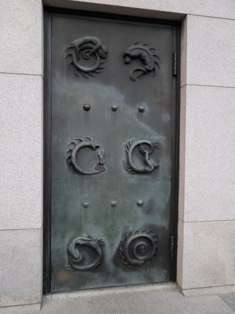 Kuva metallisesta ovesta, jossa on kuusi kohokuvaa käärmemäisestä, itsensä ympäri kiertyvästä olennosta. Joissakin on selvästi mukana myös ihminen eri elämänvaiheissaan.