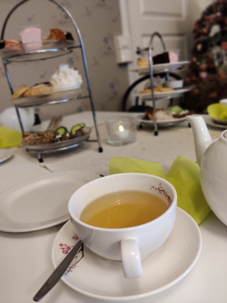 Erikoislähikuvassa kupillinen vihreää teetä valkoisessa teekupissa. Valkoinen teepannu näkyy hieman oikeassa reunassa. Taustalla pöydällä kaksi kolmikerroksista tarjoiluasetelmaa iltapäiväteen pikkusuolaista ja makeaa.