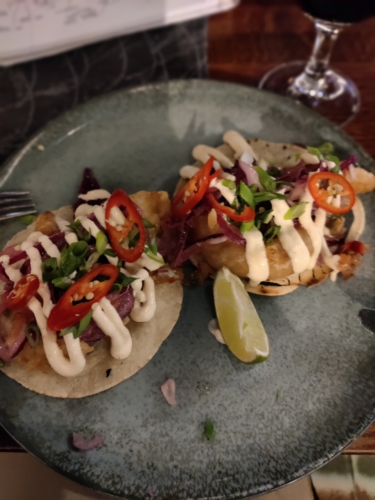 Kuvassa harmaalla lautasella kaksi pientä tacolättyä, joiden päällä kalaa, sipulia, punaista chiliä ja majoneesiraitoja. Limeviipale koristeena.