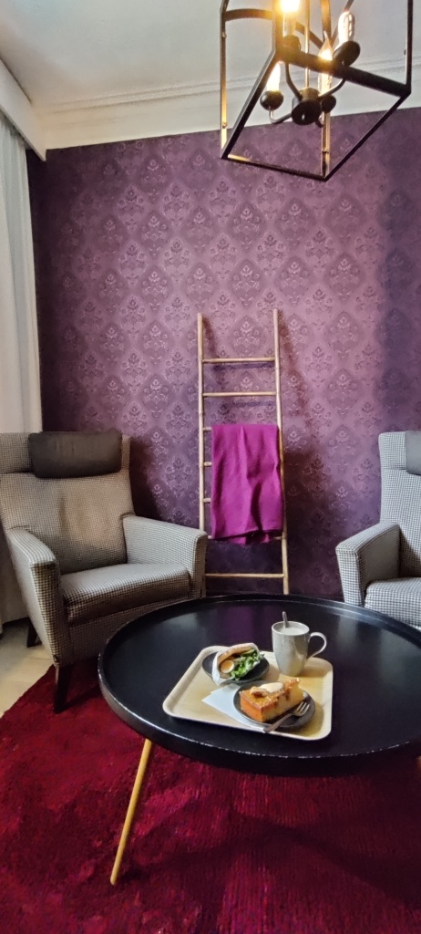 Kuva pienestä, tunnelmallisesta huoneesta, jossa on punainen matto ja matala, pyöreä pöytä. Pöydällä on tarjotin, jossa mukillinen teetä, täytetty rieska ja omenakinuskitorttu. Taustalla on kaksi nojatuolia ja koristetikkaat, joilla roikkuu violetti viltti. Seinän tapetti on violetti ja siinä on toistuvia kukkakuvioita.