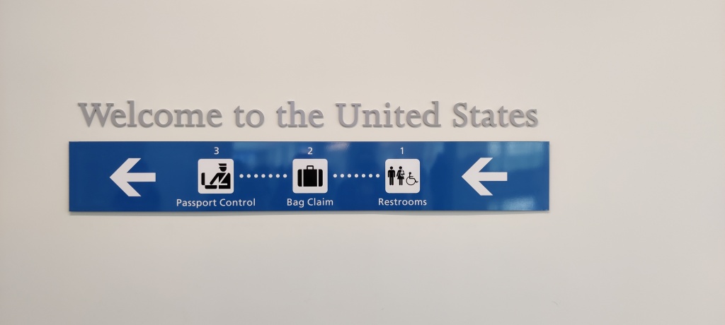 Kuvassa seinällä oleva sininen kyltti, jonka yläpuolella lukee "Welcome to the United States". Sinisessä kyltissä on kaksi nuolta, joissa kuvalliset ja sanalliset ohjeet passintarkastuksen, matkalaukkujen noutamisen ja vessojen suuntaan.